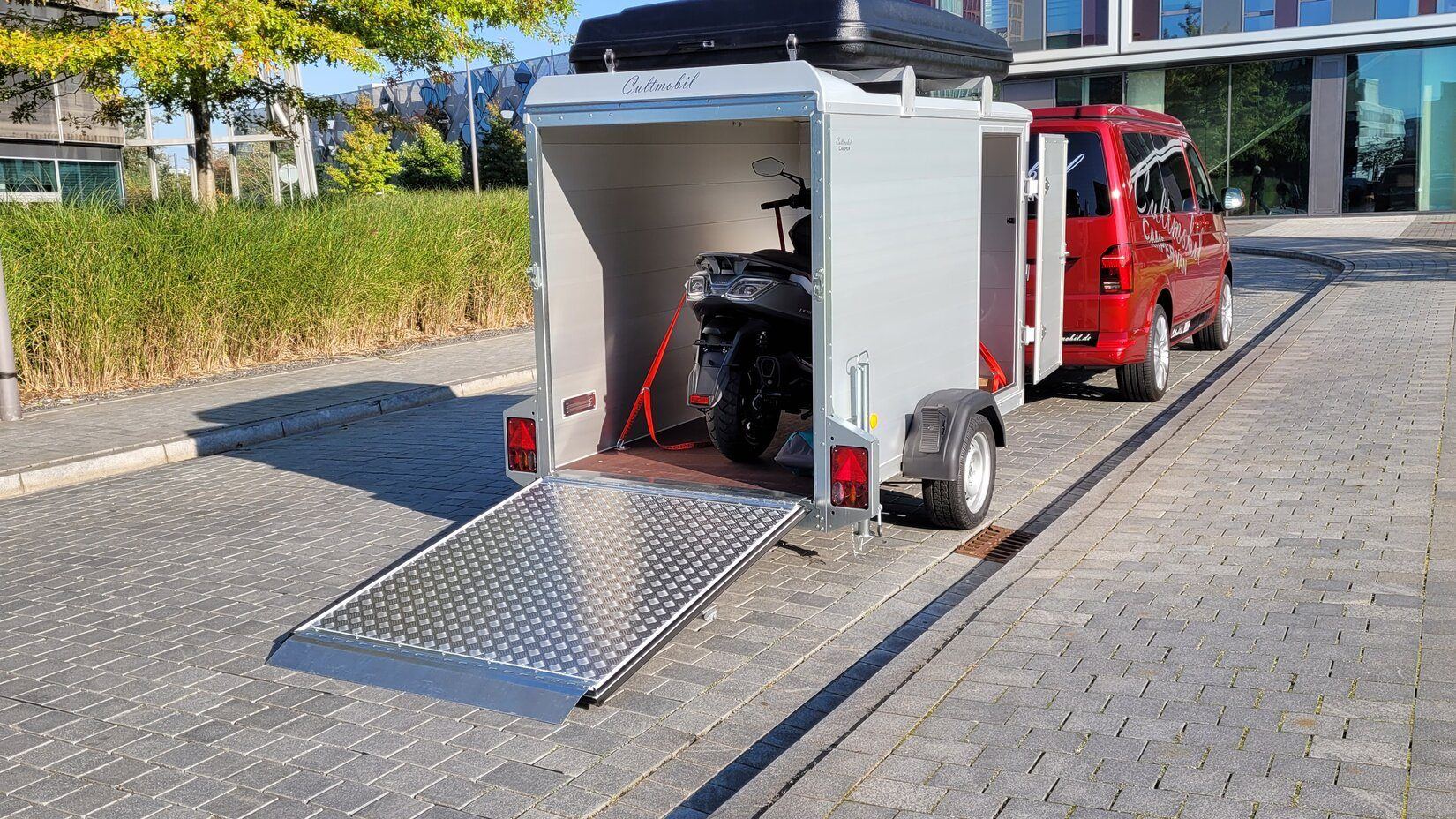 Bike Camper: geschlossener Kofferanhänger mit geöffneter Heckrampe, darin ein festgezurrter Roller, oben ein Dachzelt