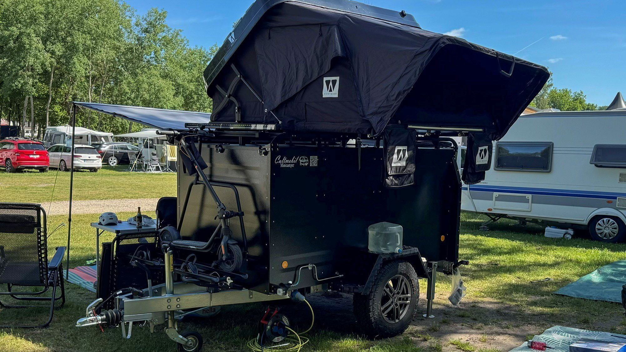 Schwarzer Dachzeltanhänger KT-EB3 Offroad Plus mit aufgeklapptem Dachzelt und Markise auf dem Campingplatz