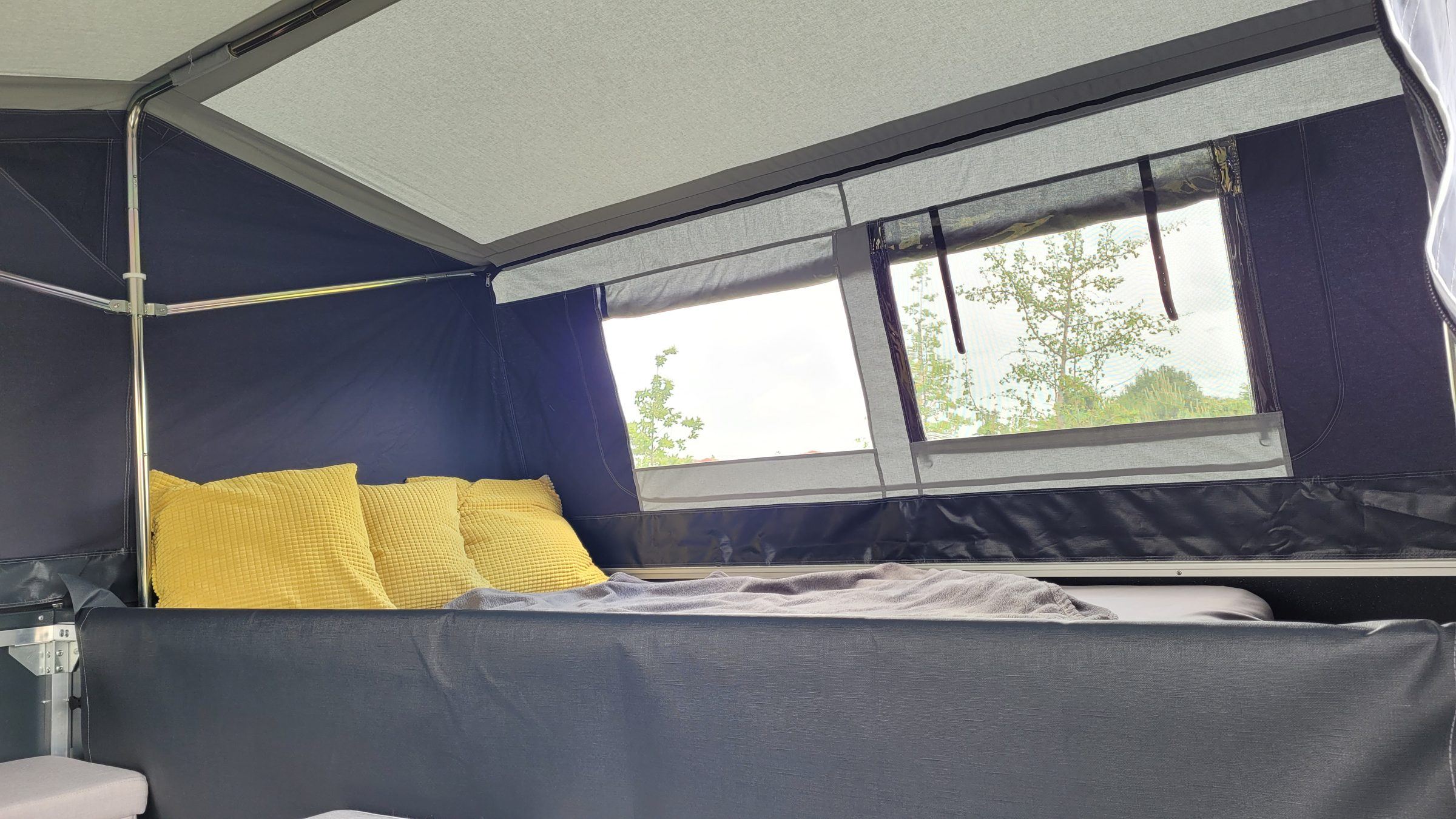 Liegefläche im Klappcamper mit Matratze, gelben Kissen und Fenster zur Wiese