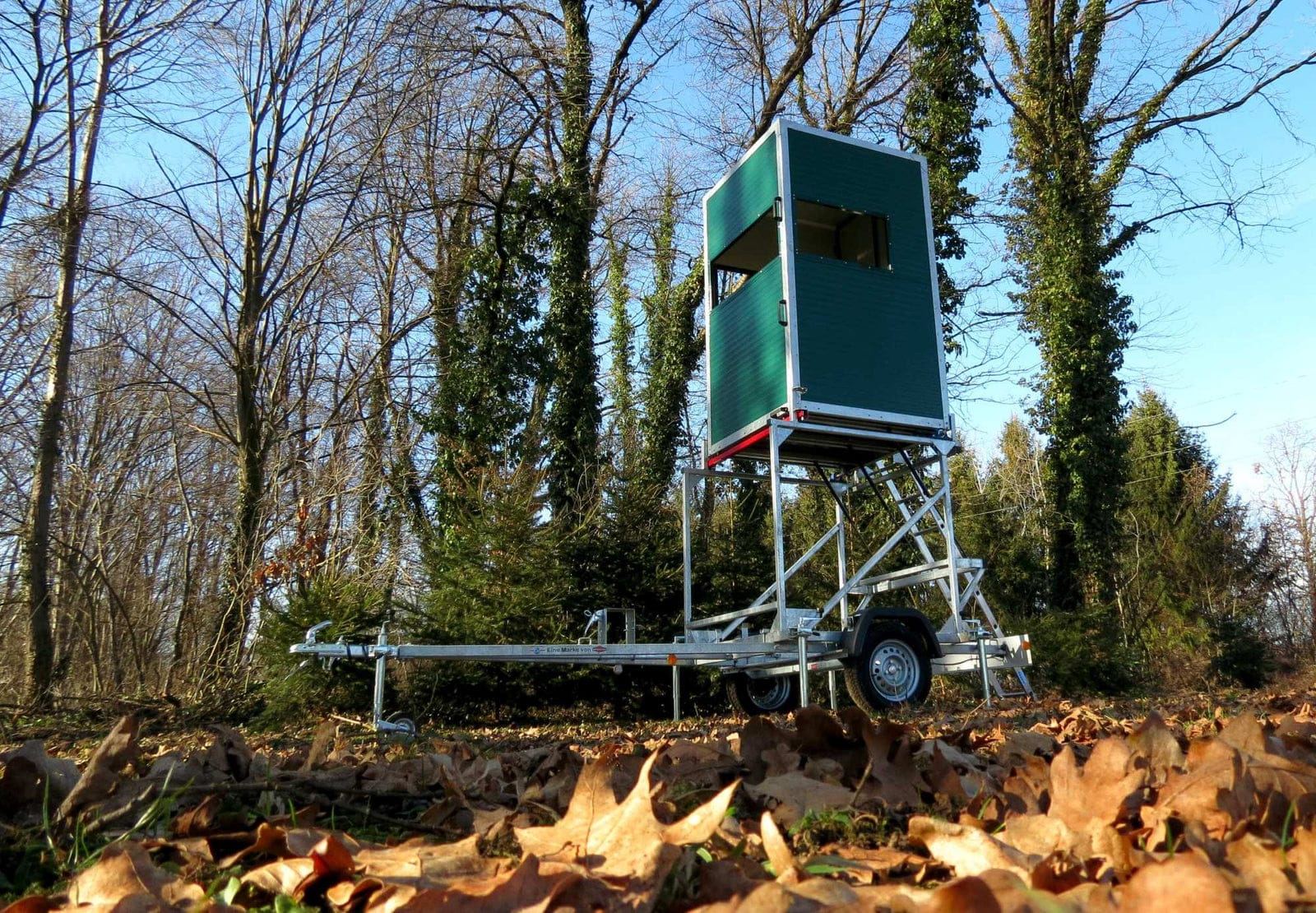 grüne mobile Jagdkanzel, aufgebaut auf einem Trailer, in der Mitte des Bildes auf Rasen, mit viel Laub im Vordergrund und kahlen Bäumen im Hintergund