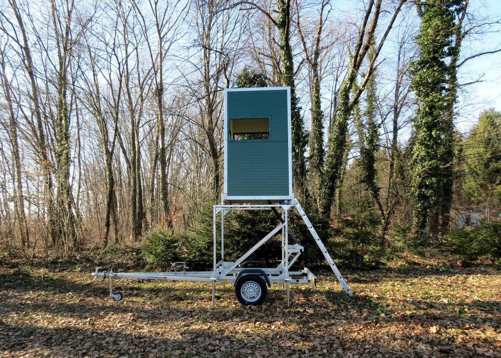 grüne mobile Jagdkanzel, aufgebaut auf einem Trailer, in der Mitte des Bildes auf Rasen, mit viel Laub im Vordergrund und kahlen Bäumen im Hintergund. Rechts lehnt eine Leiter an der Kanzel