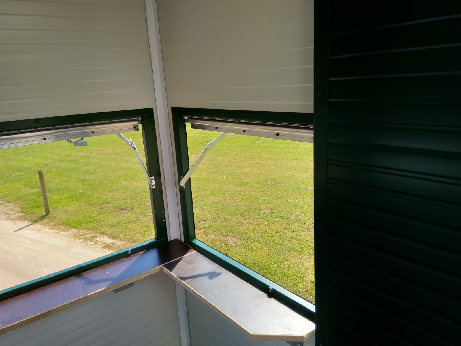 Ausblick aus einer grünen Jagdkanzel auf ein Stück grünen Rasen und etwas Weg. Die Fenster sind aufgeklappt