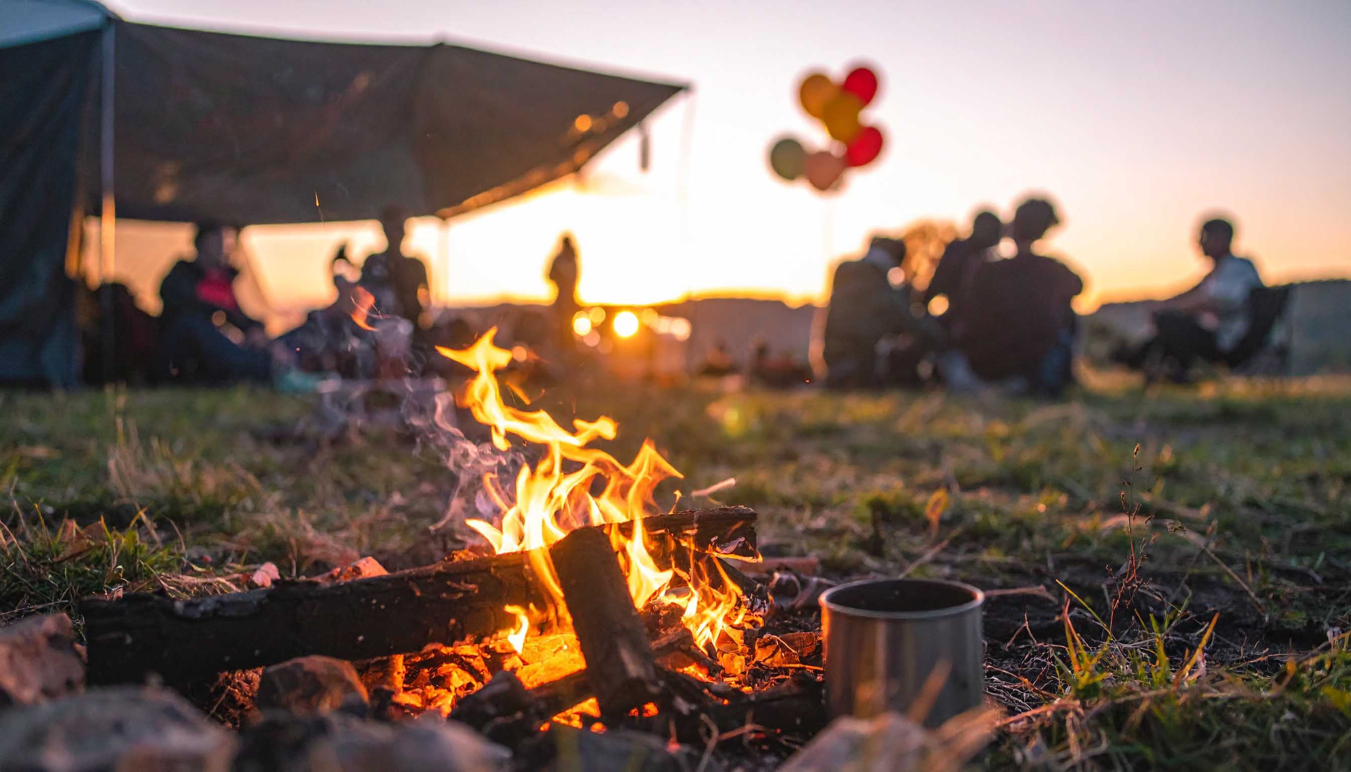Lagerfeuer am Abend, dahinter Menschen am Zelt in der Abendsonne